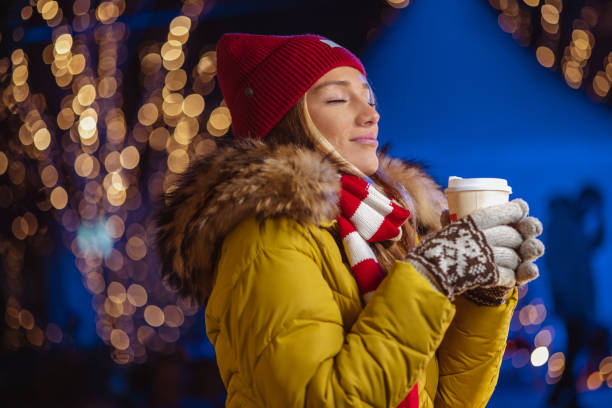Young beautiful woman with eyes closed smiling and holding warm drink. She is enjoying the magigal Christmas moments in the city