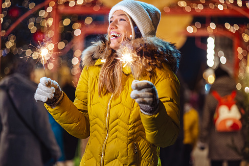 Young woman in yellow jacket holding sparklers in hands and enjoying Christmas holidays outdoors