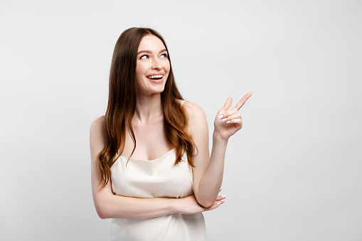 Smiling beautiful woman with long shiny hair in nightgown showing finger on copy space isolated on gray background. Happy female looking away, shopping. Concept of shopping, sale