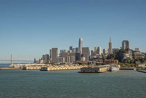 San Francisco, CA, USA - July 13, 2023: Urban jungle and skyscrapers of financial district behind docks and warehouses of port under blue sky. Oakland bridge on horizon