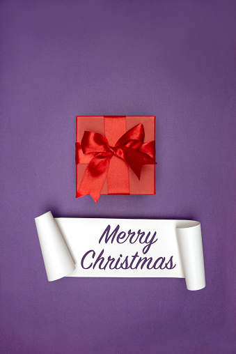 Red gift box with red ribbon on purple background and white card with Merry Christmas text.