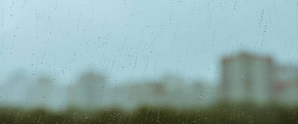 istock Atmospheric minimal backdrop with rain droplets on glass. Wet window with rainy drops and dirt spots closeup. Blurry buildings and green trees under blue sky against dirty window glass with raindrops. 1662329499