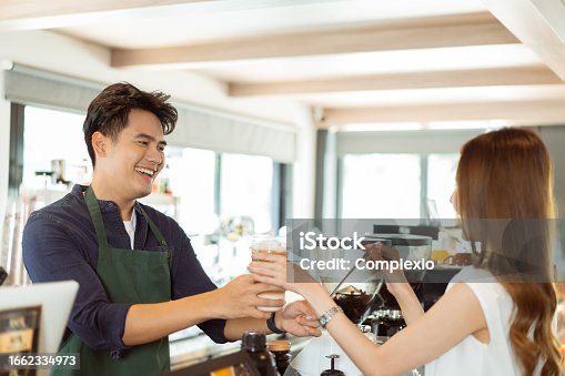 istock Asian male barista handing iced coffee to female customer over the counter at coffee shop cafe 1662334973