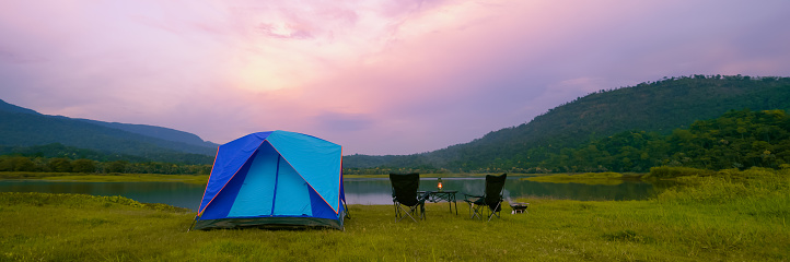 Camping Tent Near the Natural Lake and Mountain View with Outdoor Gear, Rustic Lantern on Table and Camping Chairs, Babeque grill in Evening Time. Panoramic View.