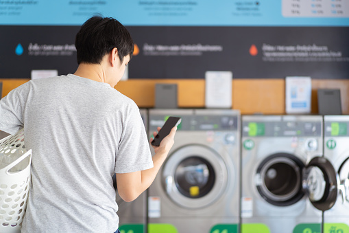 Asian man putting an used or dirty clothes in the self-service automatic laundry washing machine, Asian man using smartphone to access self service KIOS laundry machine.