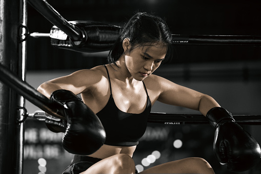 Tired young woman boxer wearing boxing gloves breathing while sitting in boxing ring.