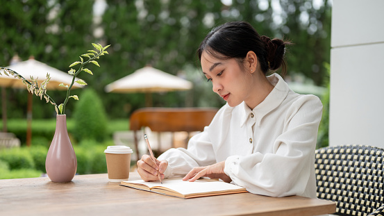A beautiful young Asian woman is writing something in her notebook, keeping a diary, or doing homework while relaxing at an outdoor cafe.