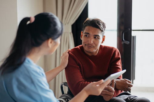 An Asian Chinese female doctor explaining medical condition report to a person with disability in a wheelchair with digital tablet during a home visit