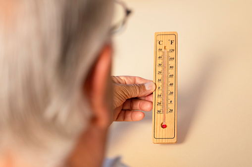 Over the shoulder view of senior man holding thermometer in hand showing high temperatures in front of beige background.