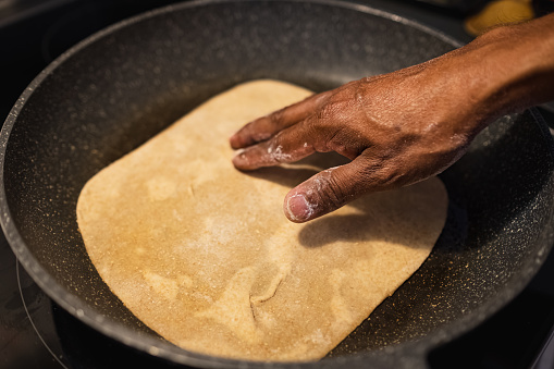 Black man frying flat bread at home. Flat bread with on the frying pan. Fresh multi-grain tortillas. Male hand and homemade naan bread on the pan