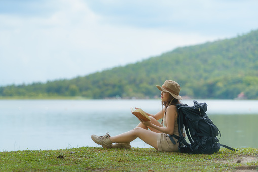 Asian female backpacker holding a book and sitting near the lake.