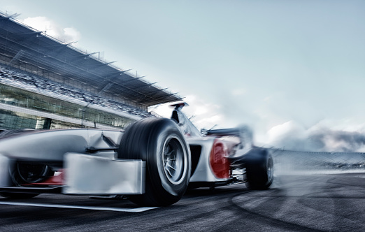 A generic red sports car viewed from the front and side as it drifts around a bend in a racetrack. The car is producing tire smoke from its back wheels and is moving fast with motion blur to the track, stand and wheels. With dramatic evening sunlight.