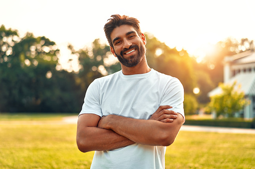 Handsome young bearded man in white t-shirt standing arms crossed in a meadow in a park on a sunny warm day, relaxing and enjoying nature on his day off.