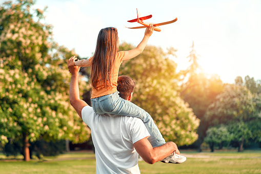 Happy father holding his little daughter on his shoulders playing with an airplane in a meadow in a park on a warm sunny day, having fun together on a day off.