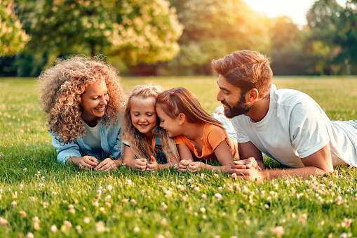 Happy young family dad, mom and two daughters lying on the grass in a meadow in the park on a warm sunny day, having fun together and relaxing on a day off.