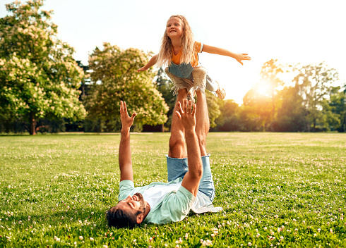 Happy young father playing with his daughter lying in a meadow in the park, holding her on his feet, having fun and relaxing on a warm sunny weekend.