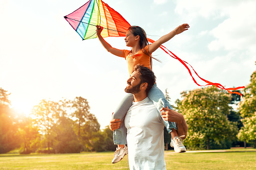 Happy young father playing with his beloved daughter with an air colorful kite in a meadow in the park, having fun together on a warm sunny weekend.