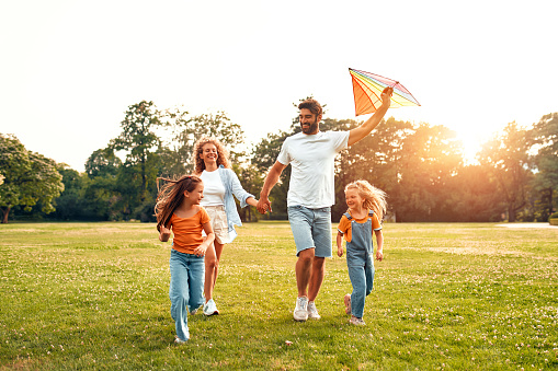 Happy young family dad, mom and two daughters running and playing with a colorful kite in a meadow in the park, having fun on a warm sunny weekend.