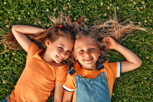 Two little sister girls lying on the grass in the meadow in the park, playing and having fun together on a warm sunny weekend.