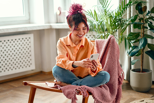 Young Asian woman with bright pink locks of hair sitting in a cozy armchair with a smartphone in the living room at home.