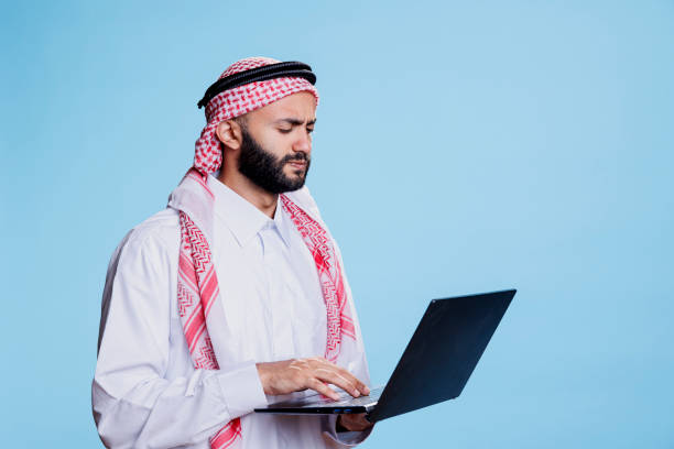 Confused muslim man working on laptop Confused muslim man wearing traditional clothes working on laptop and typing on keyboard. Arab person dressed in thobe and headscarf holding portable computer and writing message 2632 stock pictures, royalty-free photos & images