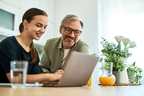 A happy father and daughter using a laptop at the kitchen table at home.