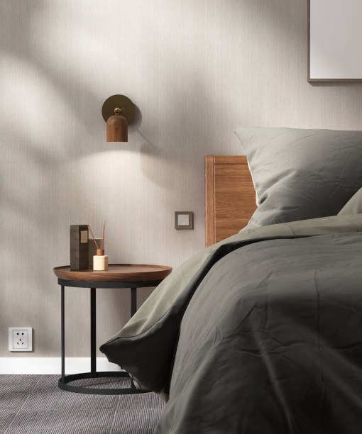 Round wooden bedside table, lamp by headboard bed, gray blanket and pillow in sunlight on vertical stripe wallpaper wall