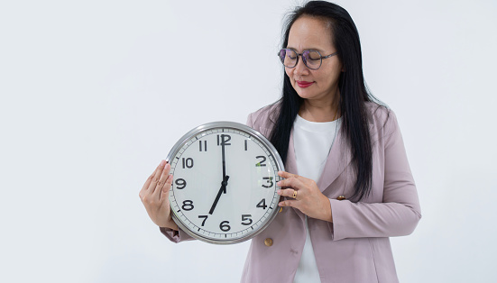 Smiling business Asian woman in eyeglasses holding big clock while looking at the clock over white background