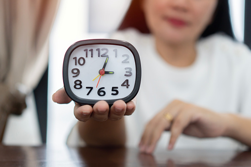 Mature woman hand holding black and white alarm clock at brown wooden table on light room background. Point of view shot. Concept of female biological time.