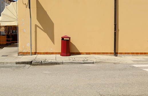 Old style red metallic street dustbin on concrete sidewalk. Light brown wall on behind and urban road in front. Background for copy space.