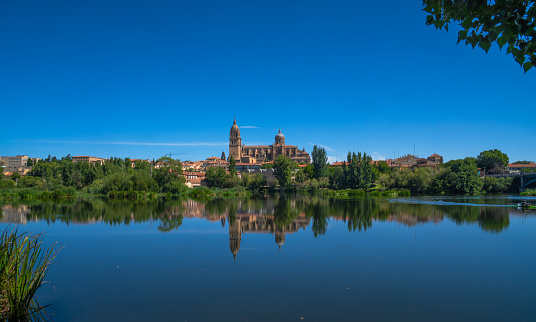 Panoramic view of Salamanca Cathedral from the other side of the Tormes River with its reflection in the calm river water and all the green vegetation, clear blue sky and clean river water.