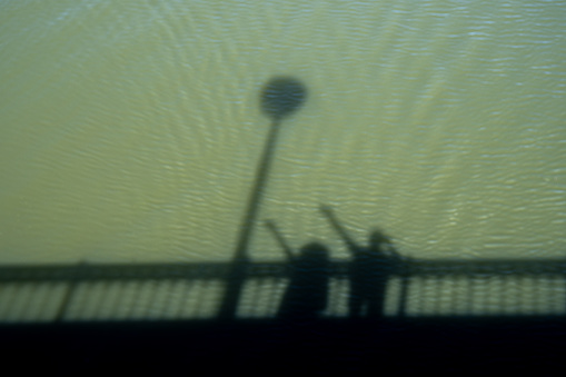 Shadow and silhouette of a bridge and a couple having a great time pointing their fingers up while taking a photo with the shadow and silhouette projected on the green waters of the Tormes River.