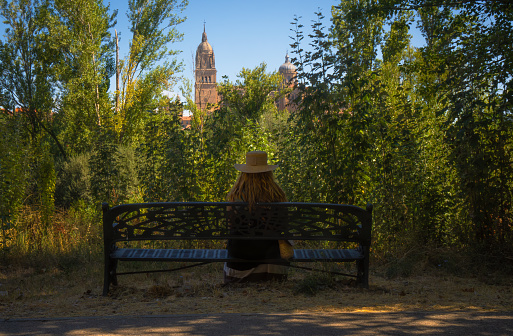 Blonde woman with a hat and straw bag and summer dress sitting on a bench with her back in a park full of trees and green vegetation looking at the Salamanca Cathedral from the other side of the river