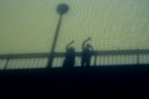Shadow and silhouette of a bridge and a couple having a great time waving while taking a photo with the shadow and silhouette projected on the green waters of the Tormes River in Salamanca.