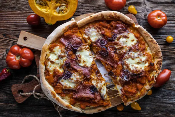 Delicious homemade pizza with italian salami, chunky pumpkin sauce, tomatoes, red onions and melted mozzarella cheese. Served ready to eat with cross section view on rustic and wooden table background. Top view