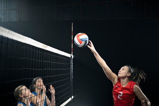 Beautiful female volleyball player performs an emotional game moment on the dark foggy background with net while holding a ball. She is wearing an unbranded sports cloth.