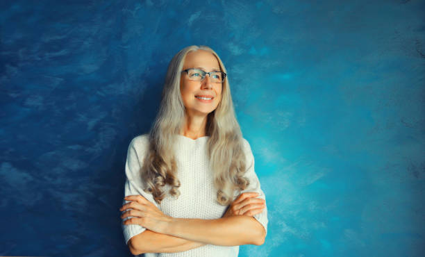 Portrait of happy smiling confident middle-aged woman with crossed arms in eyeglasses looks away. Gray-haired female in casual on blue background stock photo