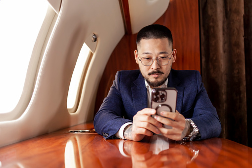 Asian successful businessman is flying in private luxury jet and using smartphone, Korean entrepreneur in suit and glasses is typing on phone in airplane