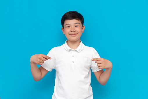 little asian boy in white t-shirt points to himself on blue background, korean child advertises copy space on his chest on isolated background