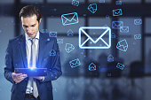 Attractive europeaqn businessman using tablet with glowing email letter icons on blurry office interior background. Communication and business message concept.