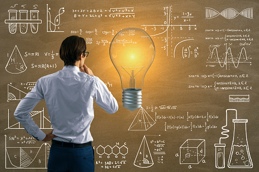 Back view of attractive thoughtful young european businessman with glowing light bulb on chalkboard wall background with mathematical formulas. Science, idea and education background