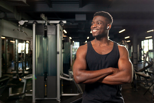 young African American man stands with crossed arms in dark gym and smiles, fitness trainer stands in fitness club and advertises copy space