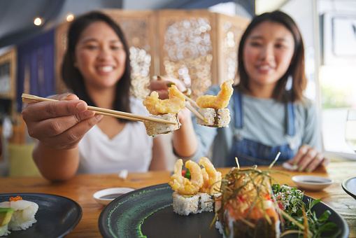 Chopsticks, girl friends and shrimp sushi at a table with salmon and Japanese cuisine food at restaurant. Young women, eating and tempura prawn with fish for lunch and meal on a plate with a smile
