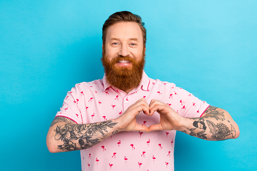 Portrait of positive satisfied man with cool tattoo wear flamingo t-shirt showing heart symbol on chest isolated on blue color background.