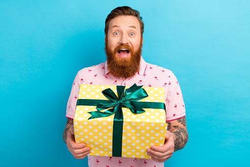 Portrait of ecstatic overjoyed man with cool tattoo wear flamingo t-shirt holding present box open mouth isolated on blue color background.