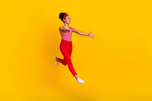 黄色の背景に興奮した小さな美しい女の子がラッシュアームをジャンプする全身プロフィール写真は、空のスペースをキャッチします - pin up girl ストックフォトと画像