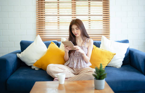 istock Happy young Asian woman sitting on sofa in living room using a digital tablet while relaxing on a sofa at home. Happy lifestyle with digital technology concept. 1663110310