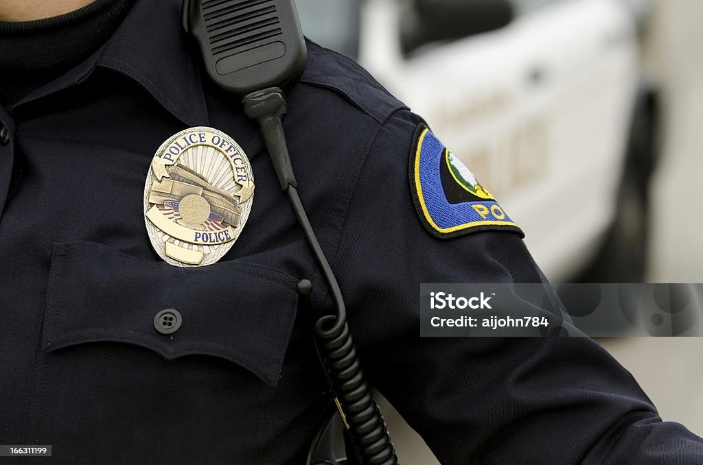 A police officer in uniform with a badge a close up of an officers uniform and badge with a patrol car in the background. Police Badge Stock Photo