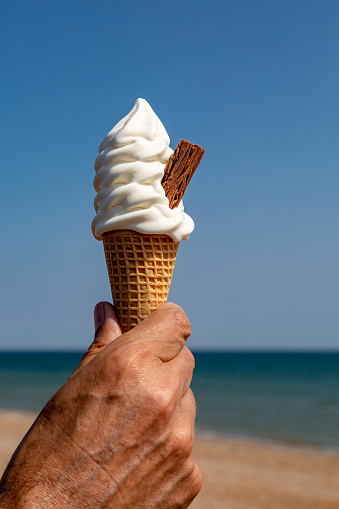 An ice cream cone being held on a summer's day, with a blue sky and the beach behind