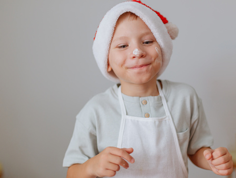A cute child in a Santa hat and with an apron, stained his nose with flour, is pleased that he will prepare a treat for Christmas.